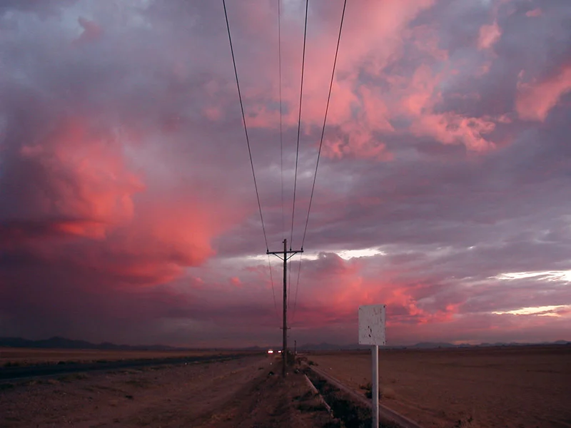 A desert road at sunset, a power line running to the vanishing point, the whole sky filled with pink and grey cloud.