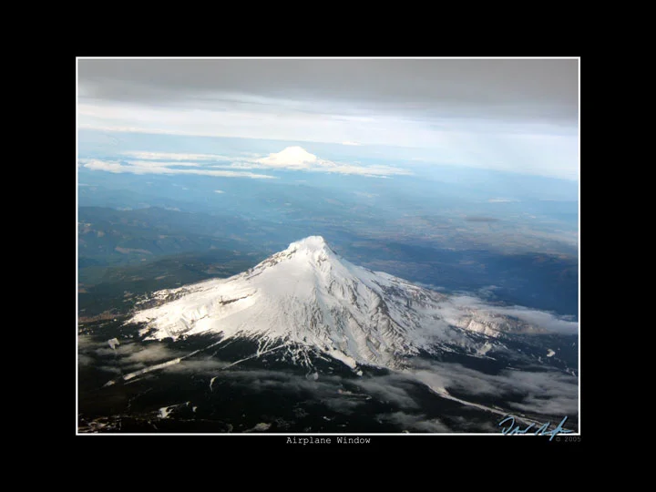 A snow-covered volcanic peak seen from an airliner window, with a thin layer of cloud below and the horizon curving away.